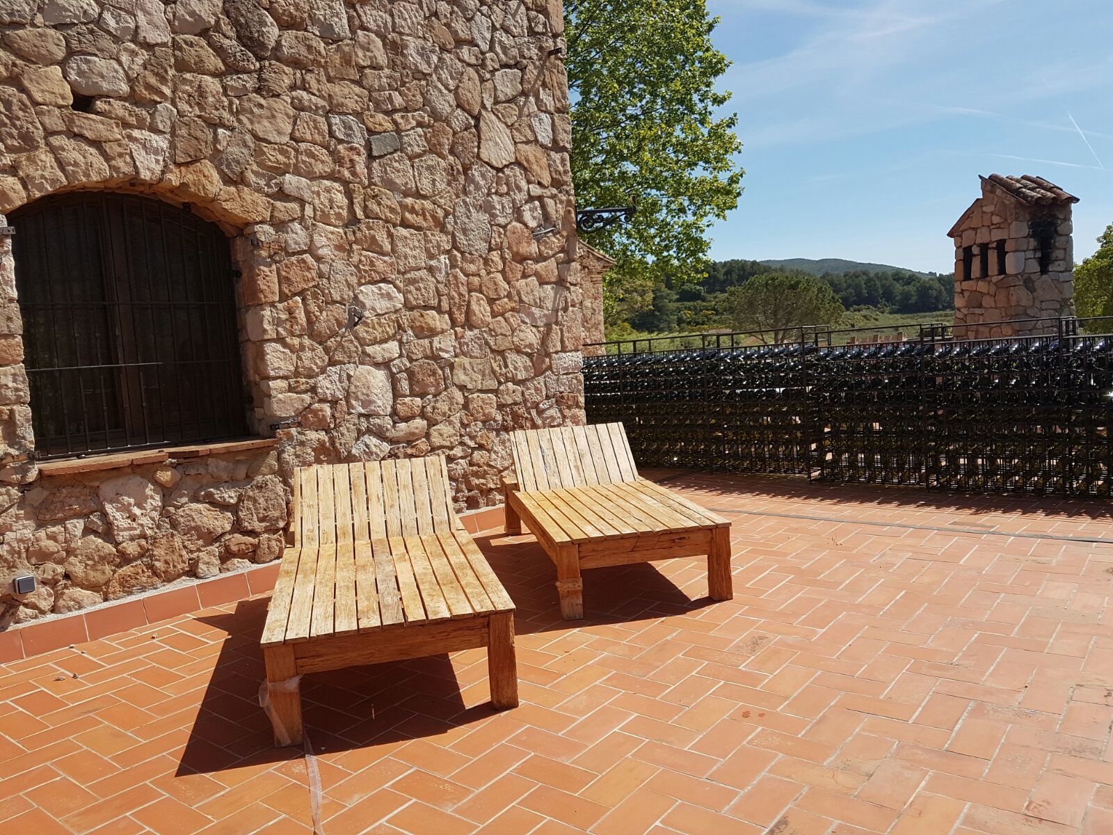 Een stenen gebouw met een groot rondboogvenster en een stenen muur. Op een betegeld terras staan twee houten lounge stoelen.