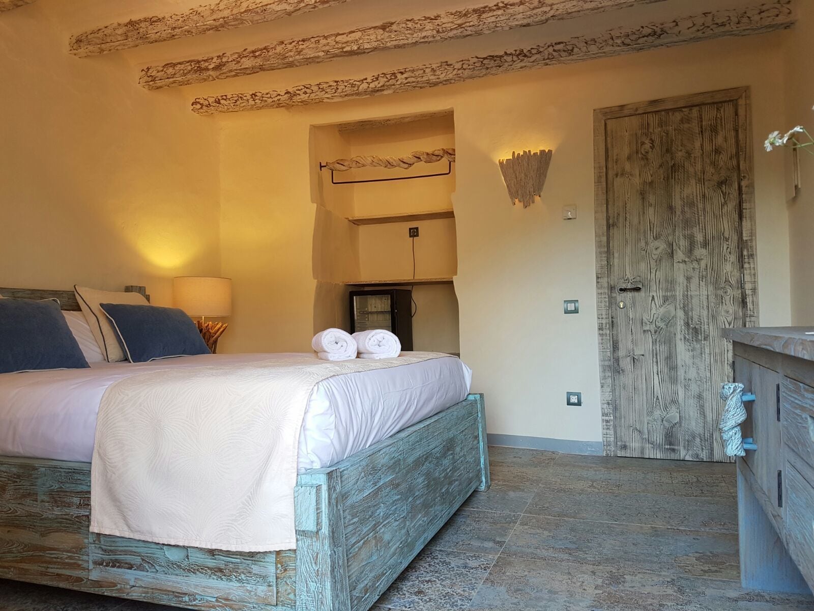 A bedroom with a bed, a wooden dresser, a door, and a wall-mounted light fixture.