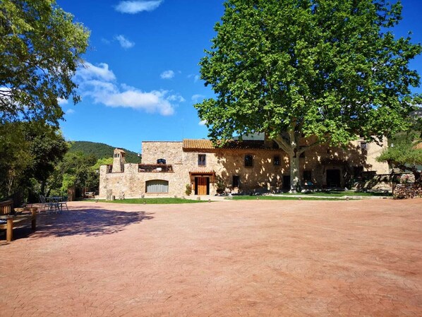 A stone building with a large tree in front, a paved courtyard, and a mountain in the background. - Mas del Sord 1335 (L'Albiol)