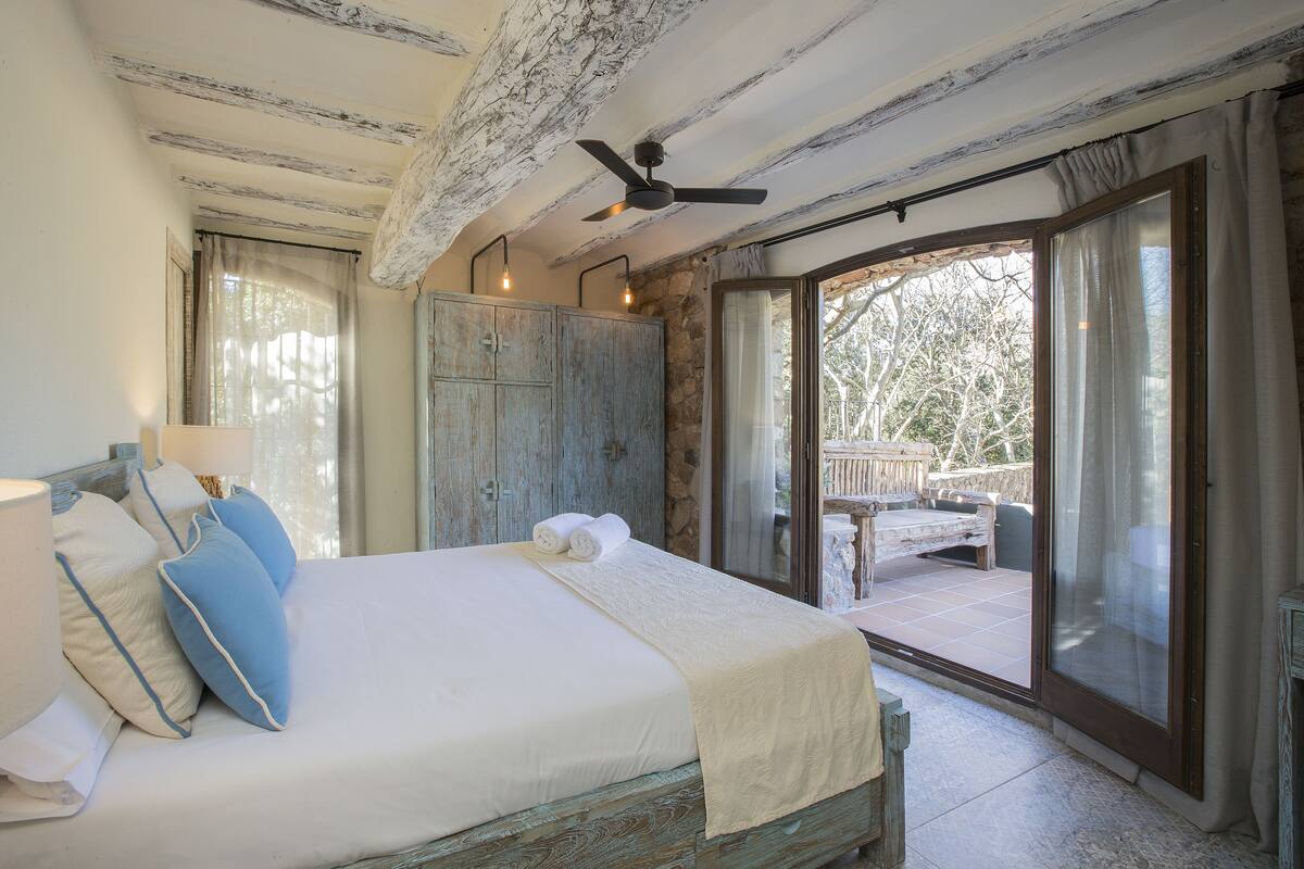 A bedroom with a bed, a ceiling fan, a wooden wardrobe, and a balcony with a table and chairs.