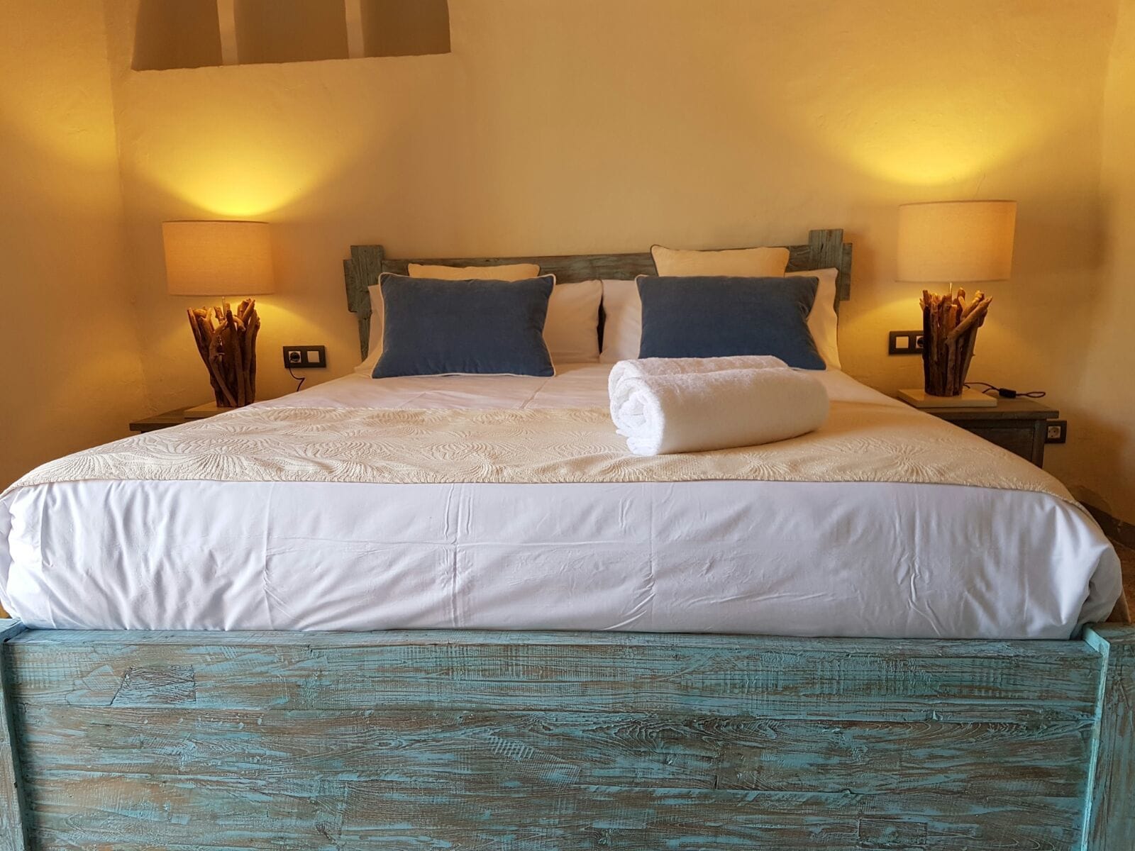 A neatly made bed with a white duvet, two pillows, and a folded towel. There are two bedside tables with lamps and decorative branches.