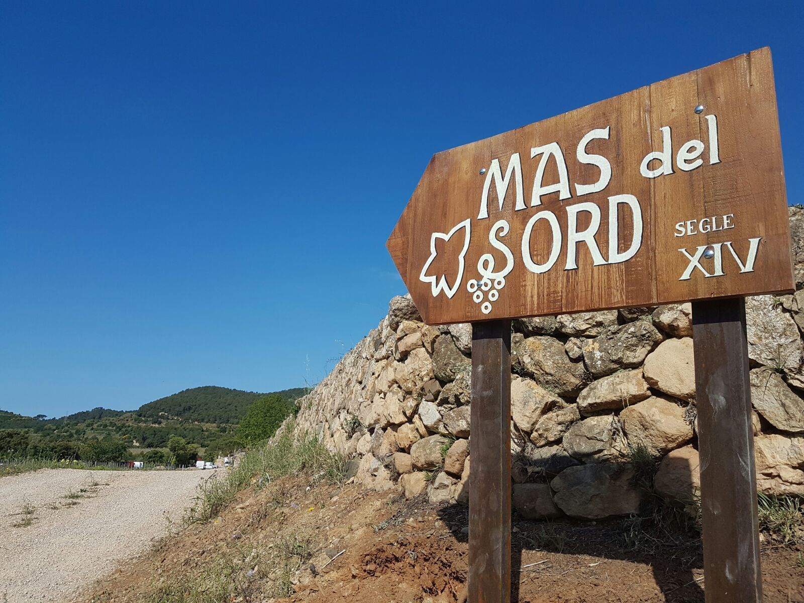 Un letrero de madera con el texto "MAS del SORD SEGLE XIV" sobre un cielo azul claro.