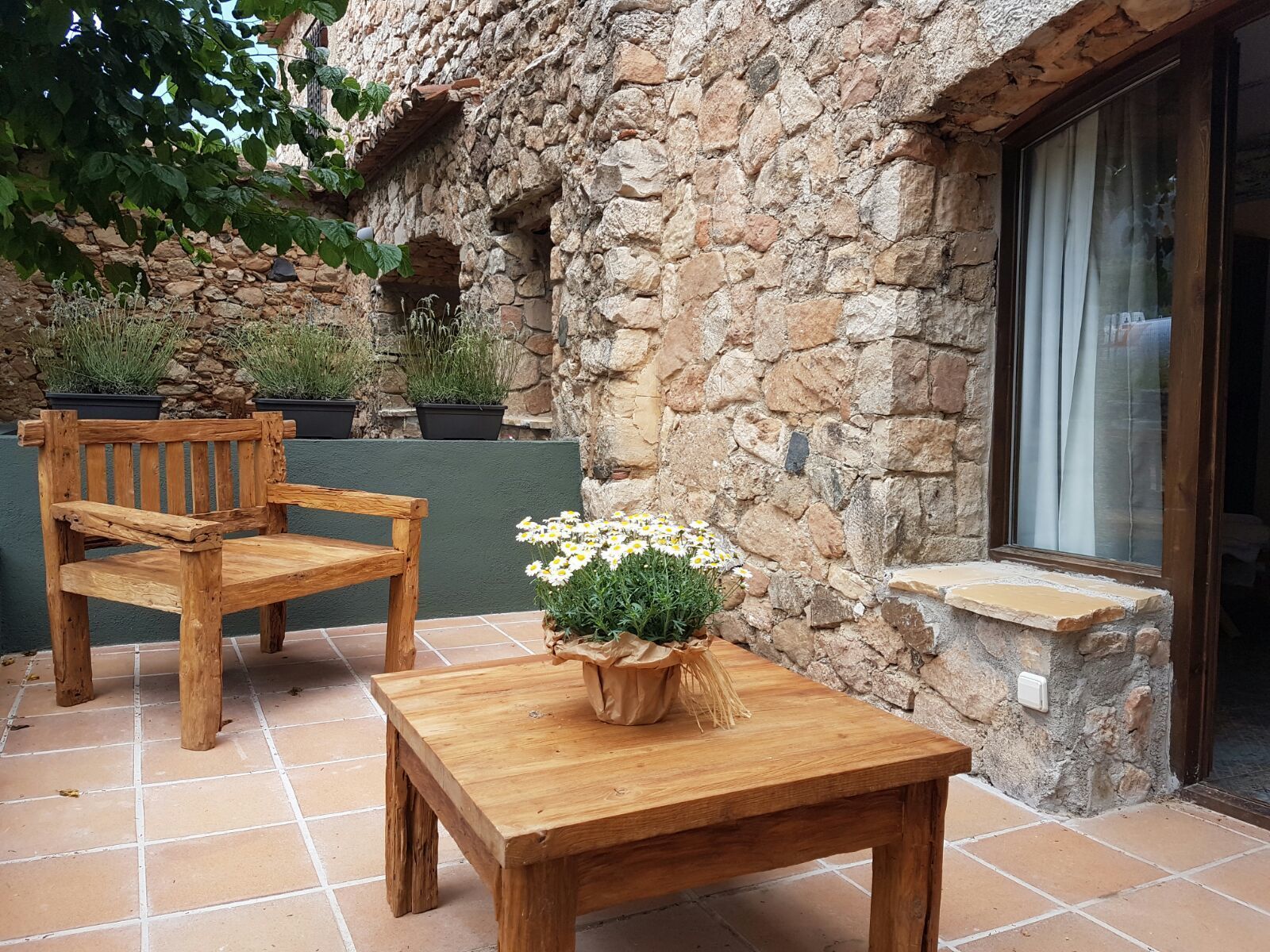 A wooden bench and table set on a tiled patio with a stone wall and potted plants.