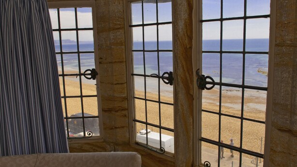A room with large windows overlooking a beach, featuring a stone wall and a curtain. - Sundial House Beachfront Arts & Crafts Period House (Lyme Regis)