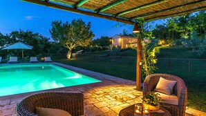A pool area with a pergola, wicker furniture, and a well-lit house in the background. - Muffato 6, Emma Villas (Sarteano)