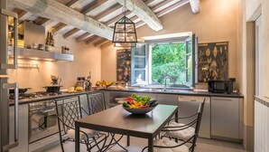 A modern kitchen with a dining table, a bowl of fruit, and a window with a view of greenery. - Muffato 6, Emma Villas (Sarteano)
