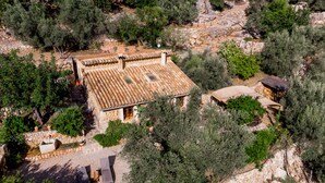 Una casa de piedra con tejado de baldosas, rodeada de olivos y un paisaje rocoso.