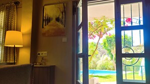 Puerta abierta que da a un jardín con piscina, una lámpara sobre una mesa y un cuadro en la pared.