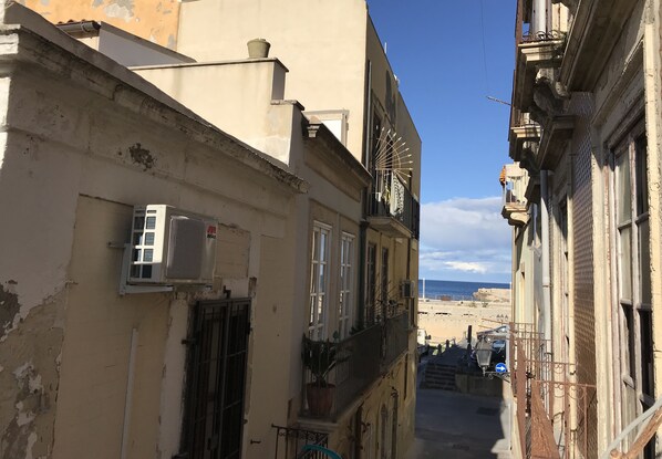 A narrow street with buildings on either side, a visible air conditioning unit, and a view of the sea in the distance.