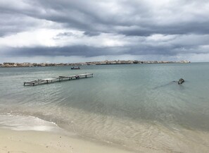 A coastal scene with a pier extending into calm waters, a sandy beach, and a distant cityscape under a cloudy sky.