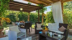 Un espacio exterior techado con mobiliario de mimbre, una mesa con cesta de fruta y una mesa de vidrio con velas.