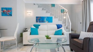 Een moderne woonkamer met een witte bank, een glazen salontafel en een bruine fauteuil. Achtergrond toont een trap met een witte leuning.