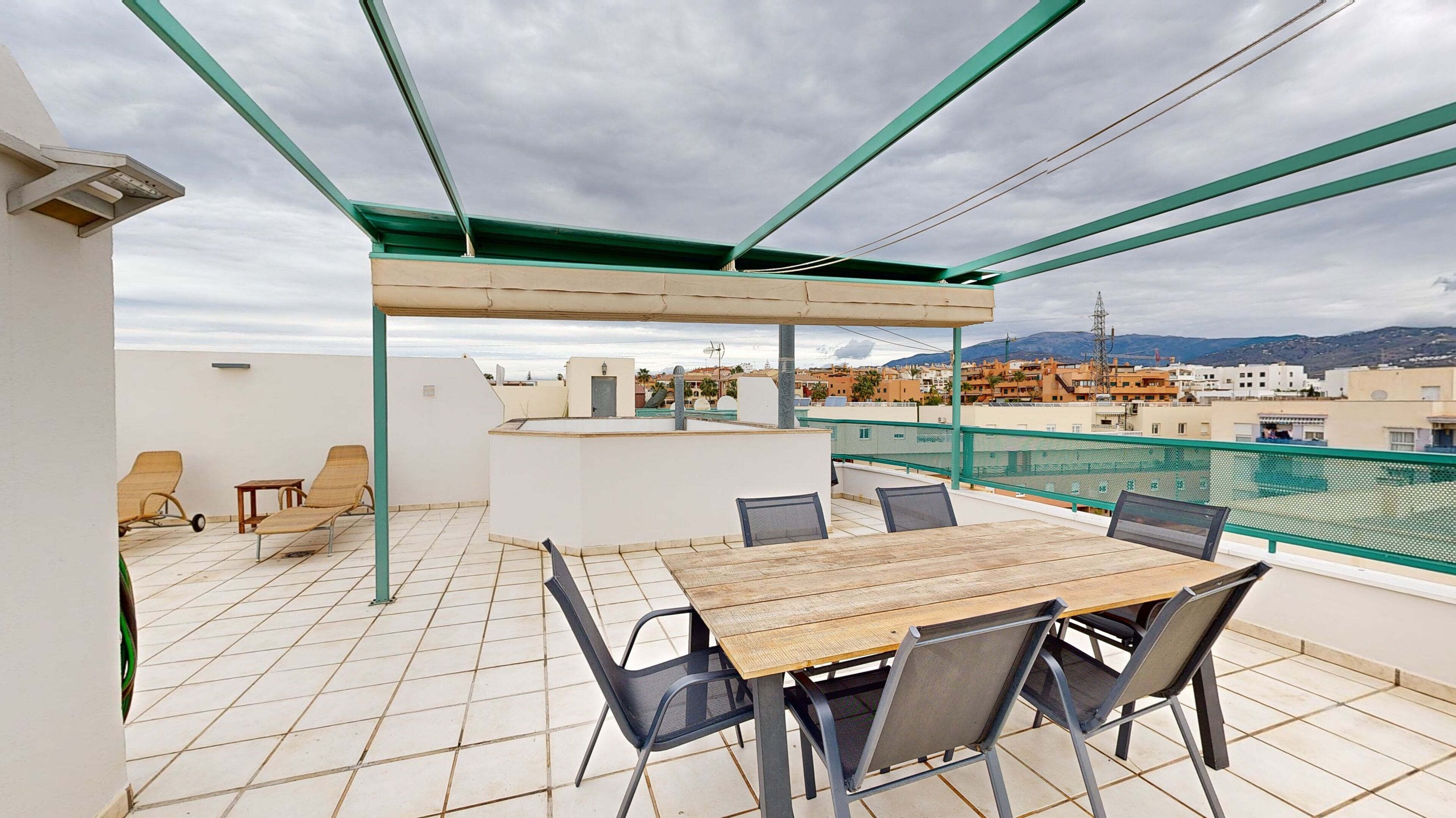 Eine Dachterrasse mit Holztisch und -stühlen, einem überdachten Sitzbereich und Blick auf die Stadt.