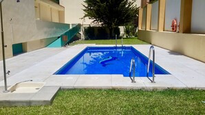 A rectangular swimming pool with a blue mosaic design, surrounded by a concrete deck and a grassy area.