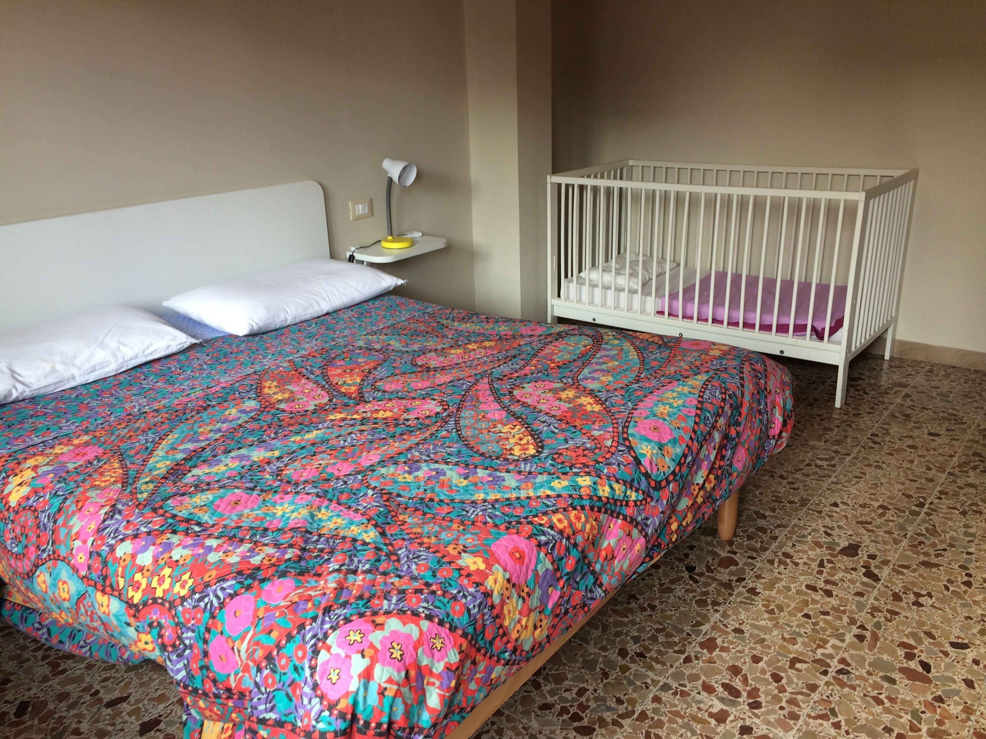 Et soveværelse med et farverigt sengetæppe, en hvid vugge og et natbord med en lampe.