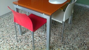 Taula de menjador amb un plat blanc i un suport per plats negre, envoltada de cadires amb coixins vermells i grocs.