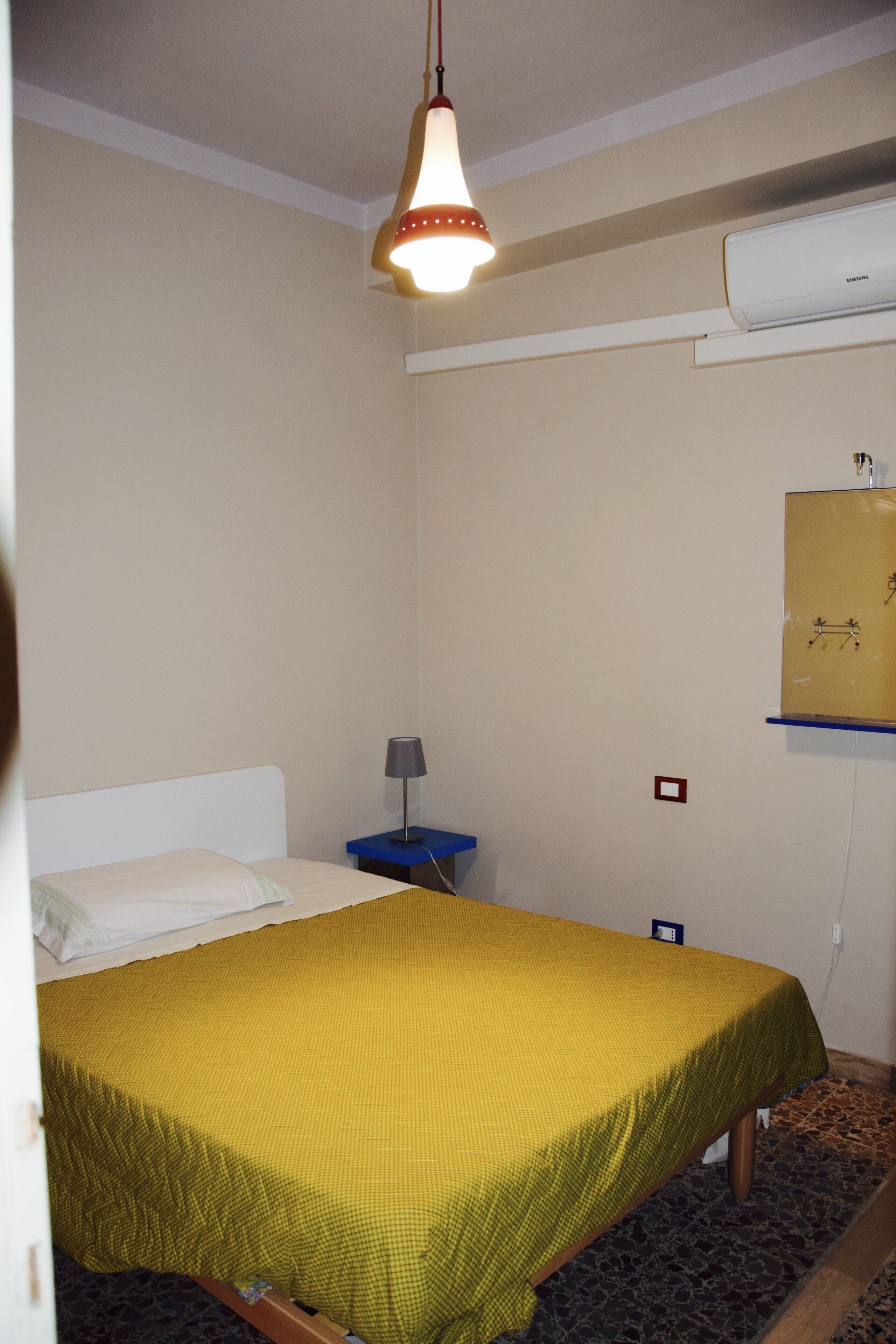 A room with a bed, a nightstand, a lamp, an air conditioner, and a wall-mounted shelf.