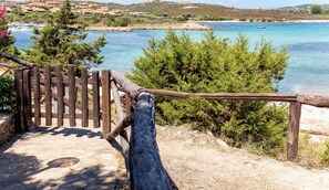Eine Holztreppe führt zu einem Strand mit klarem, blauem Wasser und Blick auf eine Küstenstadt im Hintergrund.