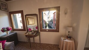 Una stanza accogliente con soffitto in legno, una finestra vista su una stretta via, un tavolino con una lampada e un divano con cuscini.