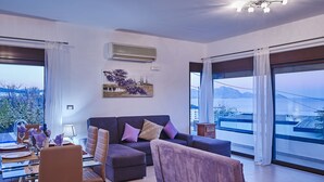 Uma sala de estar moderna com sofá, cadeiras, uma mesa de jantar posta para dois e grandes janelas com vista para um corpo d'água e edifícios ao longe.
