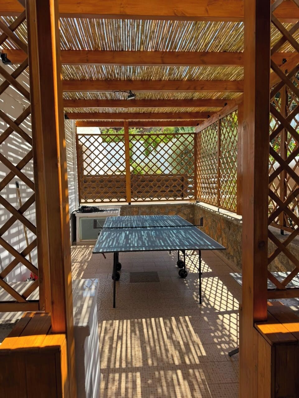 An outdoor ping pong table under a wooden pergola with a lattice design, surrounded by a stone wall.