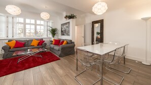 A modern living room with a glass-top dining table, grey sofas with colorful cushions, a red rug, and pendant lighting. - London House, Chiswick, Luxurious, 3 Large Beds - 4 x Bath - GREAT PLACE TO STAY (London)