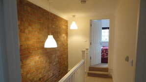 A hallway with a brick wall, white railing, and a carpeted staircase leading to a room with a red bedspread. - London House, Chiswick, Luxurious, 3 Large Beds - 4 x Bath - GREAT PLACE TO STAY (London)