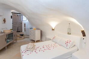 Une chambre à coucher avec un lit blanc, une commode, une coiffeuse et une salle de bain visible à travers une porte en arc de cercle.