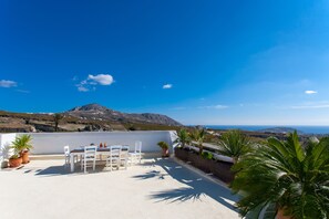 Une terrasse sur le toit, avec une table et des chaises blanches, des plantes en pot et une vue sur la mer et les montagnes.