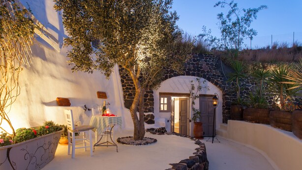 A stone-walled building with a door and windows, a potted plant, a table with a chair, and a lamp. - Spacious Luxury Private Villa - Heated Pool - Ocean Views (santorini)