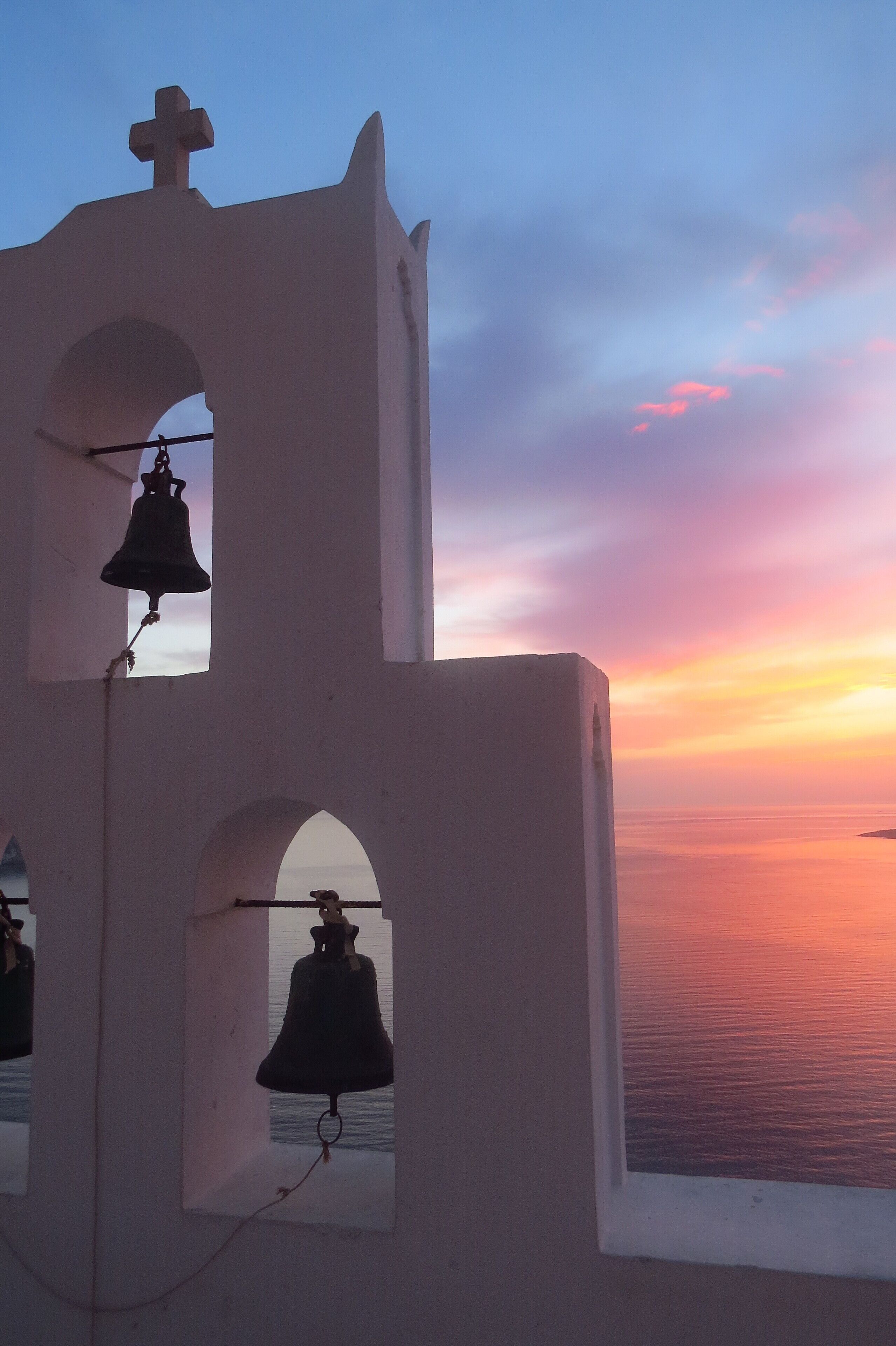 A bell tower with two bells, one inside an arch and one hanging from a balcony, overlooking a sunset over the sea.