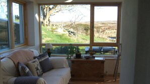 A cozy room with a large window offering a view of a grassy landscape. A wooden bench and a table with a lamp and a bowl of fruit are visible. - Traditional Failte Ireland 2025 Rustic Stone Cottage With Private River Fishing. (Clifden, Connemara)