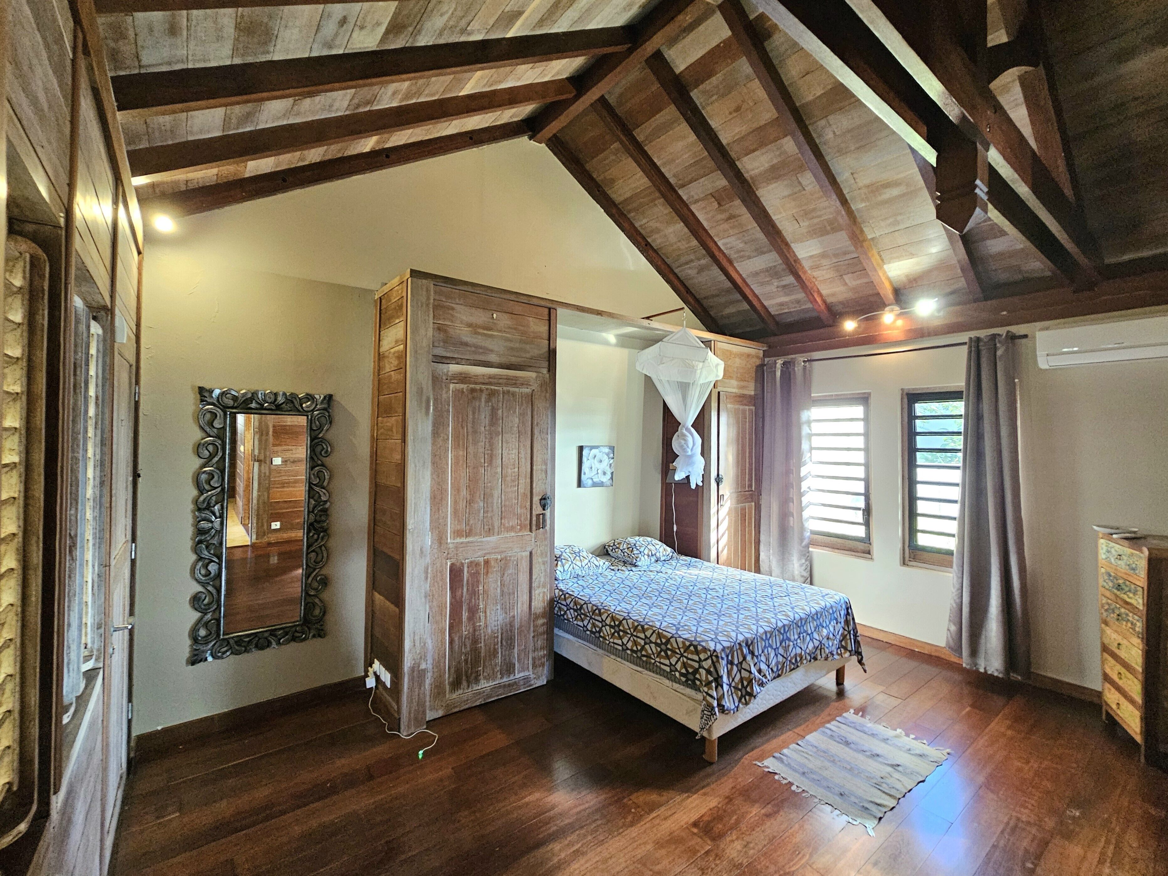 A bedroom with a wooden bed, a large mirror, a wardrobe, and a ceiling with exposed beams.