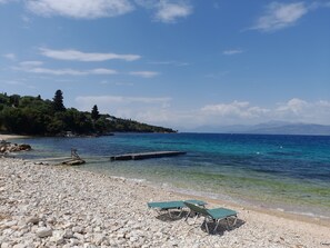 Een kiezelstrand met helder turquoise water, een houten steiger en een rij groene ligstoelen.