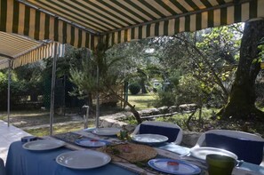An outdoor dining area with tables set under striped canopies, surrounded by greenery and a stone wall.