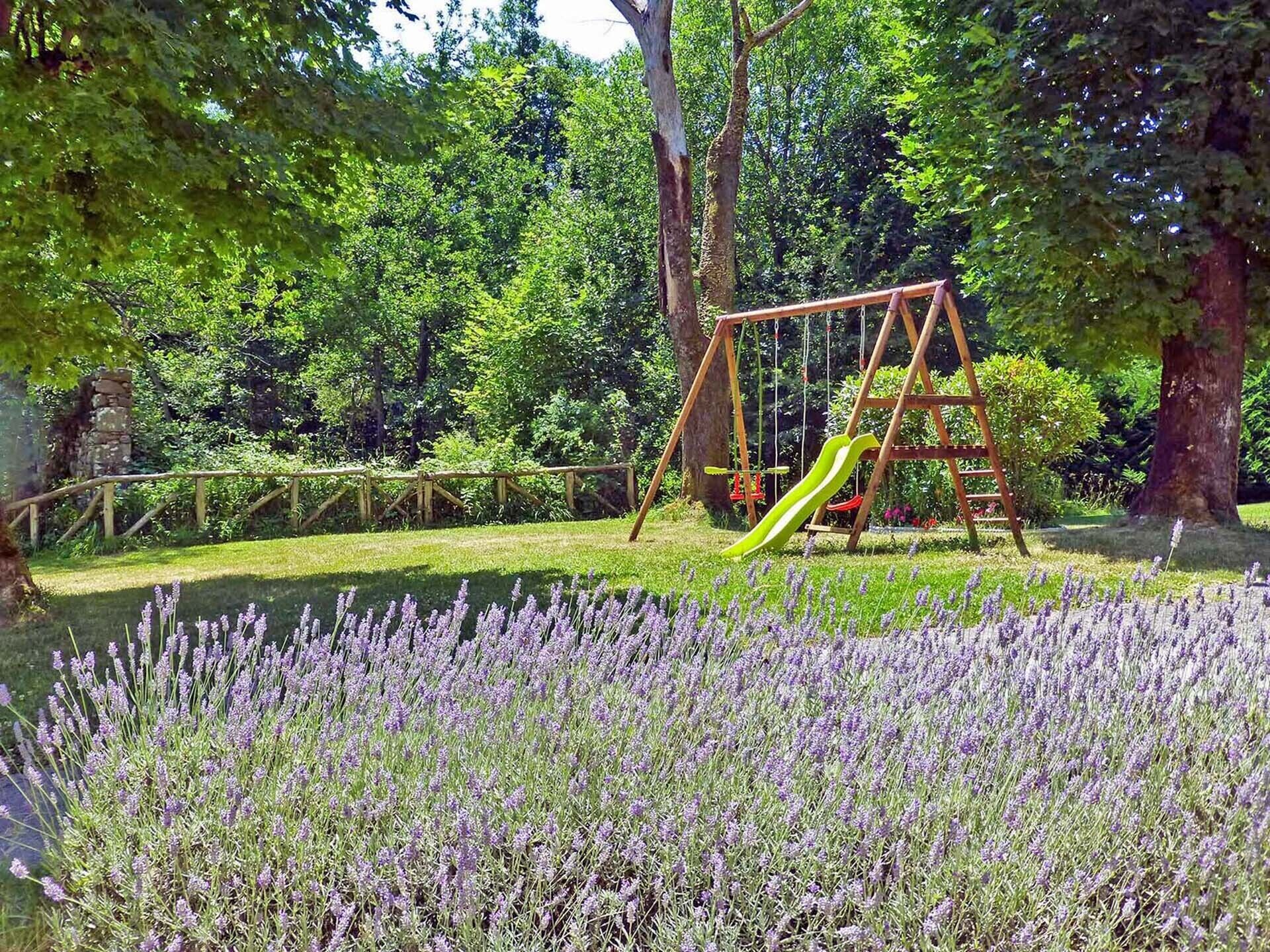 Un parque infantil con columpios y tobogán, rodeado de árboles y campos de lavanda.