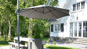 Une terrasse extérieure avec une table et des chaises, un grand parasol, et une maison en arrière-plan.