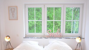 Une chambre avec un lit, deux lampes de chevet, une fenêtre donnant sur des arbres et un panneau avec le mot « Sourire » placé sur le rebord de la fenêtre.