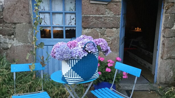Een stenen gebouw met een blauwe deur en een raam, een bord waarop “Plage du Verger” staat, en een blauwe stoel met een rieten mand vol bloemen.