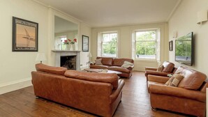 Ein geräumiges Wohnzimmer mit Ledersofas, einem Kamin, großen Fenstern und einem Fernseher.