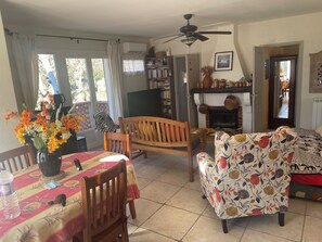 A cozy living room with a wooden sofa, a patterned armchair, a dining table with a floral tablecloth, a vase of flowers, and a fireplace.