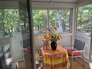 Une pièce avec une table dressée pour deux personnes, un vase rempli de fleurs et une vue sur une forêt, visible à travers de grandes fenêtres.