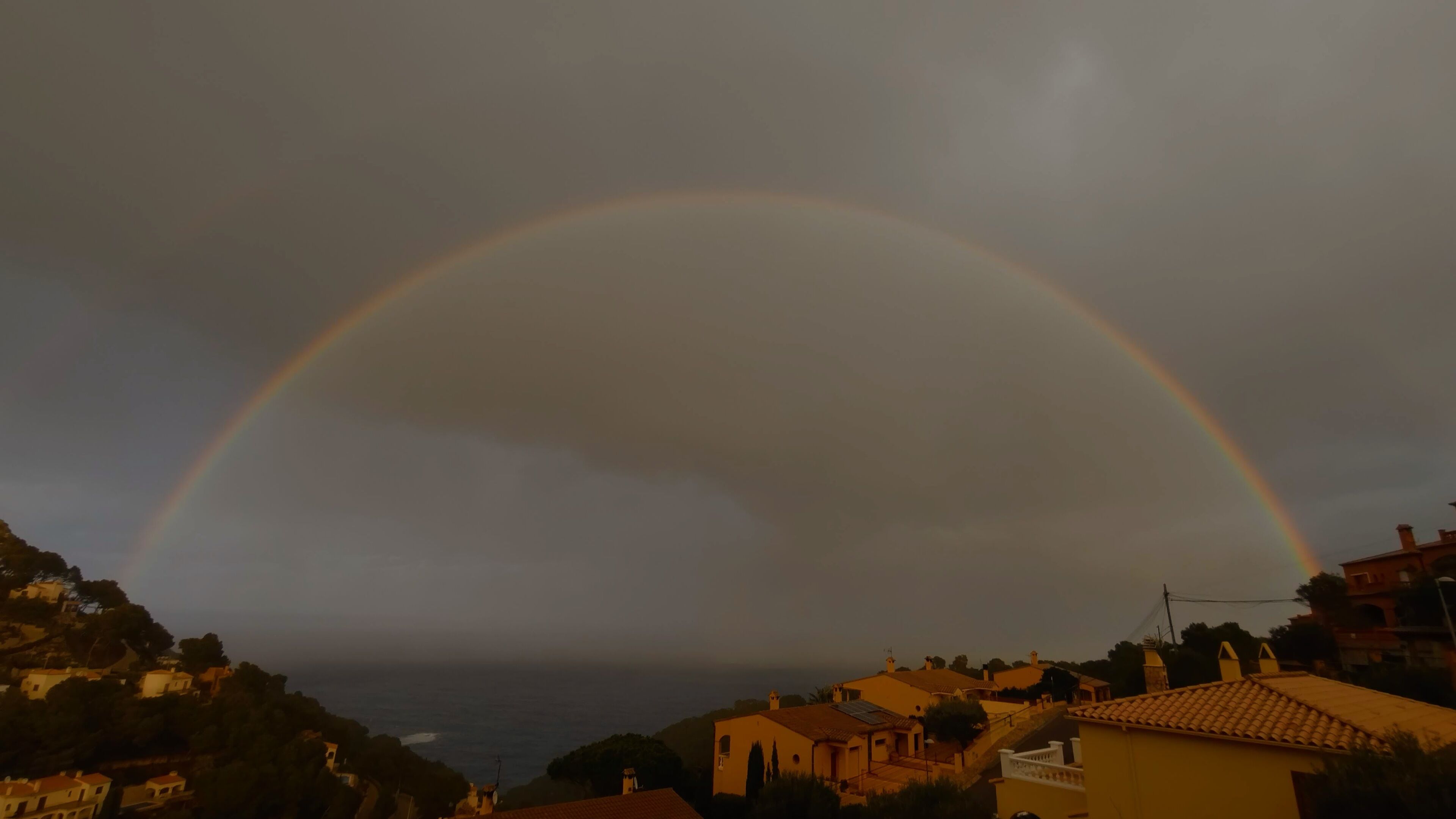 Een dubbele regenboog boven een kustplaats met huizen en de zee op de achtergrond.