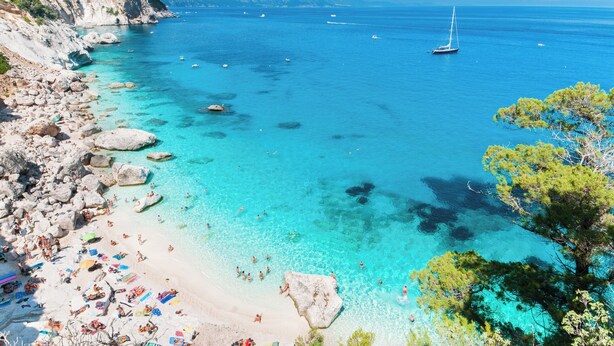Una spiaggia con acque cristalline turchesi, costa rocciosa e una barca a vela all'orizzonte.