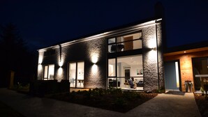 Ein modernes, zweistöckiges Haus mit großen Fenstern, gut ausgeleuchteter Fassade und einem einsehbaren Innenraum mit Küchenbereich.