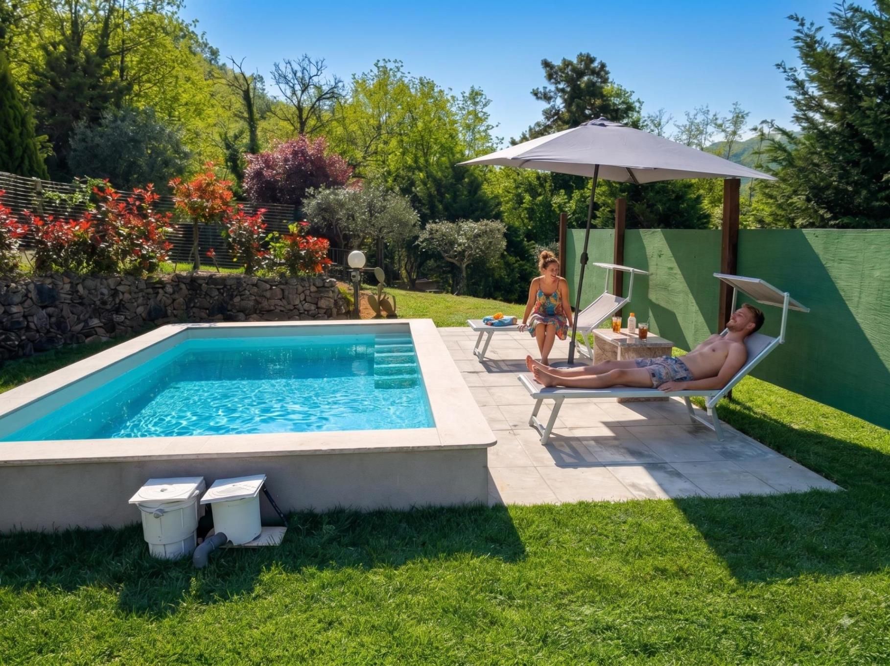 Une piscine rectangulaire bordée de pierres, entourée d’une pelouse verdoyante et d’une clôture verte.
