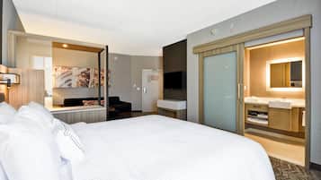 A hotel room with a large bed, a desk, a chair, a TV, a wardrobe, and a bathroom with a sink and mirror.