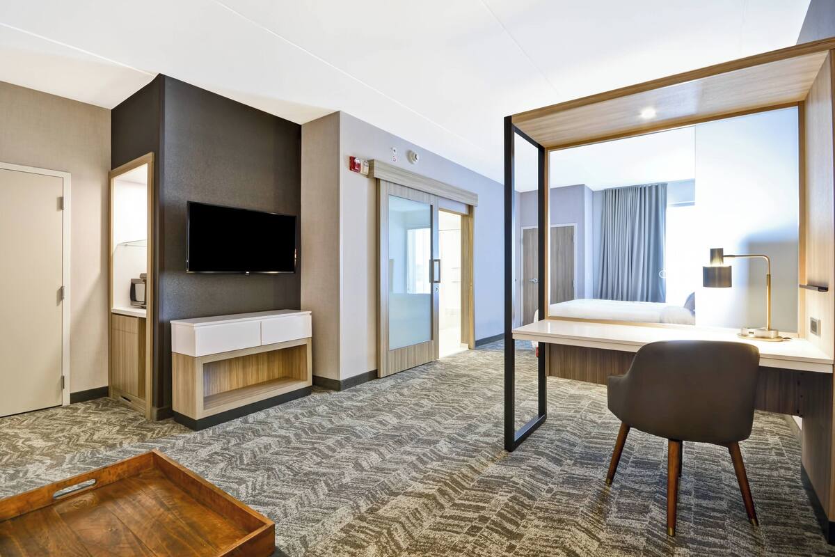 Ein Hotelzimmer mit Flachbildfernseher an der Wand, einem Bett mit großem Spiegel darüber, einem Schreibtisch mit Lampe und einer Holztablett auf dem Boden.