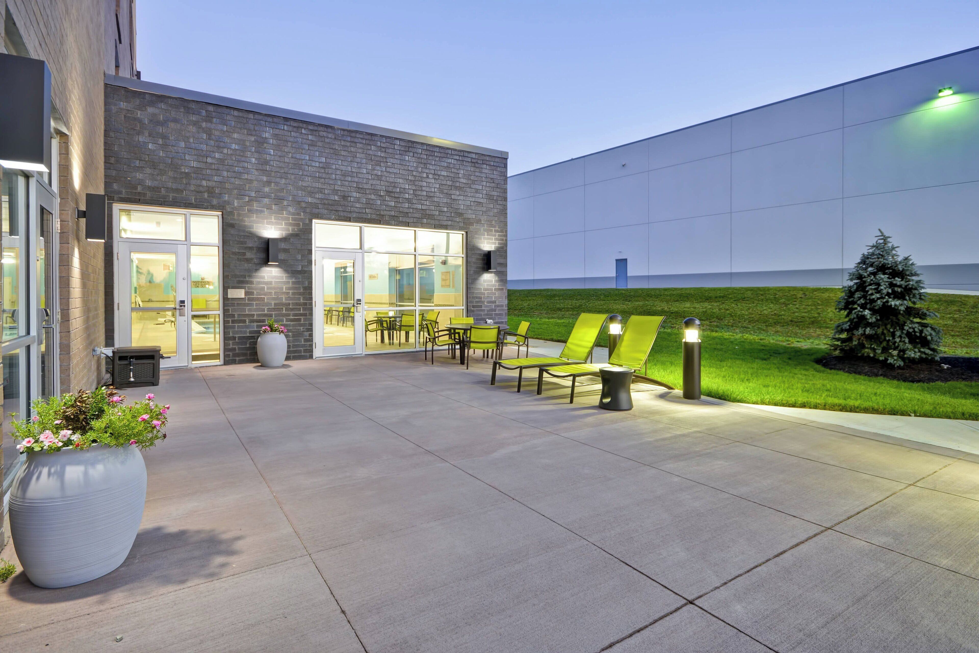An outdoor seating area with chairs and a table, a large planter with flowers, and a building with a brick facade and large windows.