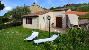 Un bâtiment en pierre avec un toit en tuiles, un petit jardin agrémenté de deux chaises longues blanches et d’un parasol rouge.
